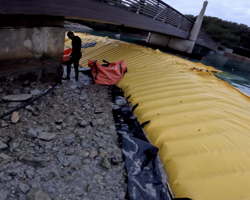 Extrémité d’un batardeau souple raccordée à une plage avec zone de travail asséchée sous une passerelle en milieu marin.